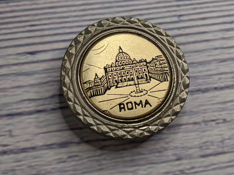 A silver and gold circle metal magnet with an engraved image of a capitol building and the word Roma.